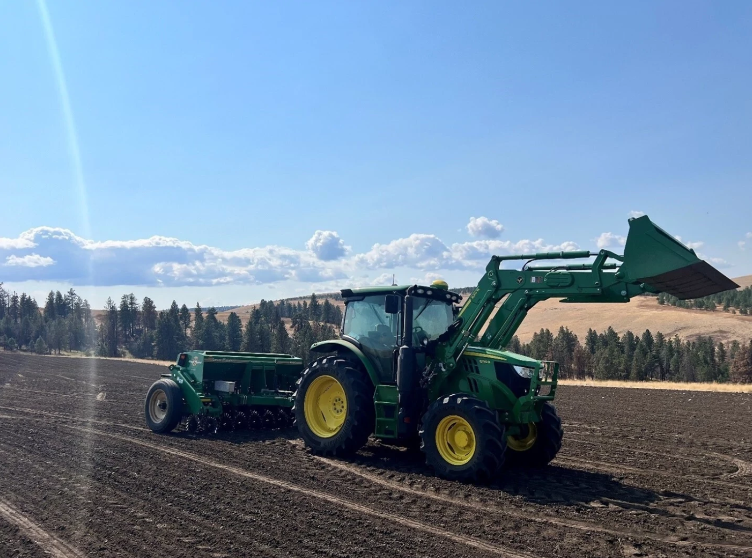 A green tractor with a front-loader attachment pulls a seeding implement across a freshly tilled field under a clear blue sky, with pine trees and rolling hills in the background.